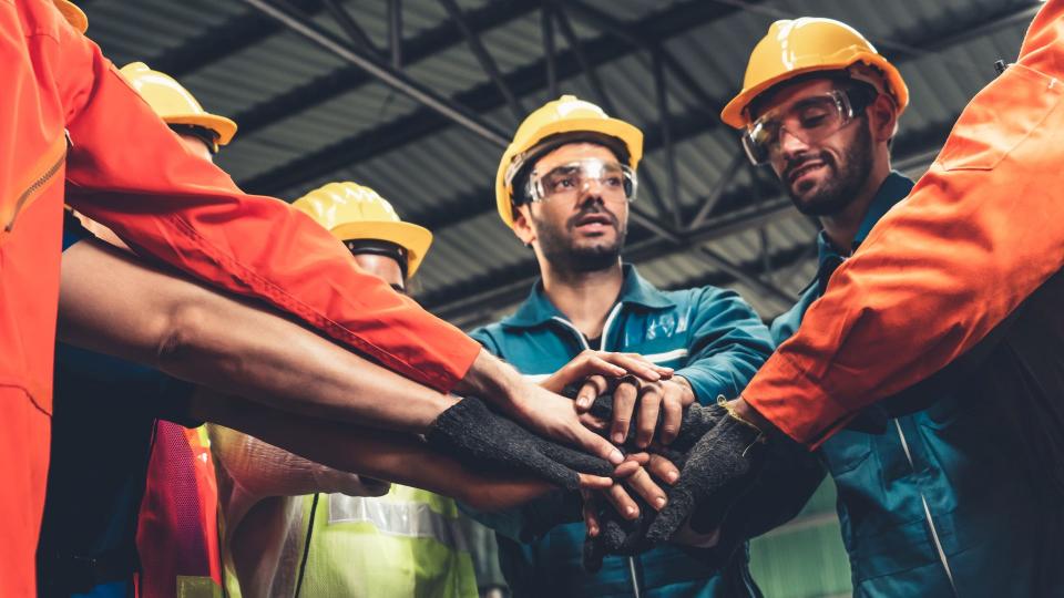 Mehrere Personen in Arbeits- bzw. Schutzkleidung stehen in einem Halbkreis zusammen und legen in der Mitte geschlossen ihre Hände übereinander. 