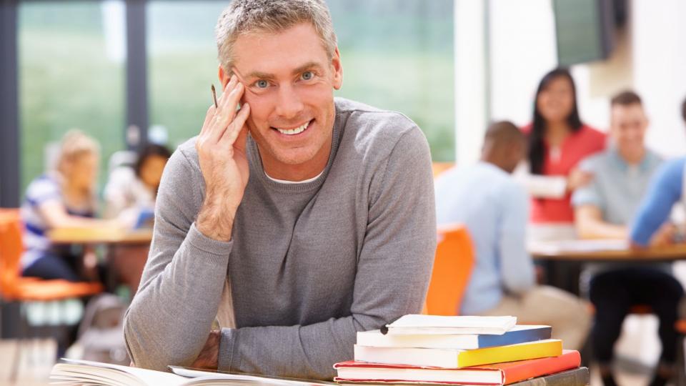 Abgebildet ist ein Mann, der lächelnd an einem Schreibtisch sitzt, auf dem sich mehrere Bücher befinden. Im Hintergrund sitzen weitere Personen an unterschiedlichen Schreibtischen. 