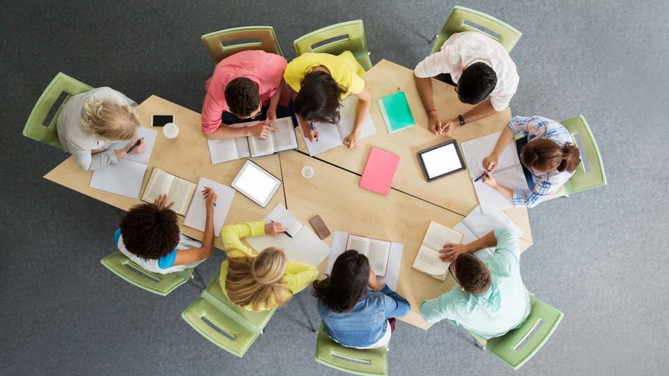 Gruppe internationaler Studenten, die mit Tablet-PC-Computern, Büchern und Notizbüchern in der Universitätsbibliothek von oben am Tisch sitzen