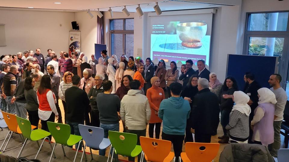 Abgebildet sind 75 Personen, die in einem Schulungsraum stehen und miteinander kommunizieren.