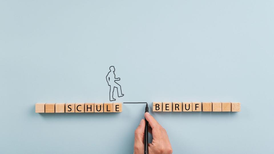 Abgebildet ist eine Hand, die eine Linie zwischen mehrere aneinandergereihte Holzwürfel zieht. Auf den linken Würfeln steht das Wort "Schule" und auf den rechten das Wort "Beruf". Eine gezeichnete Person bewegt sich von dem Wort "Schule" mithilfe der Linie zum Wort "Beruf".