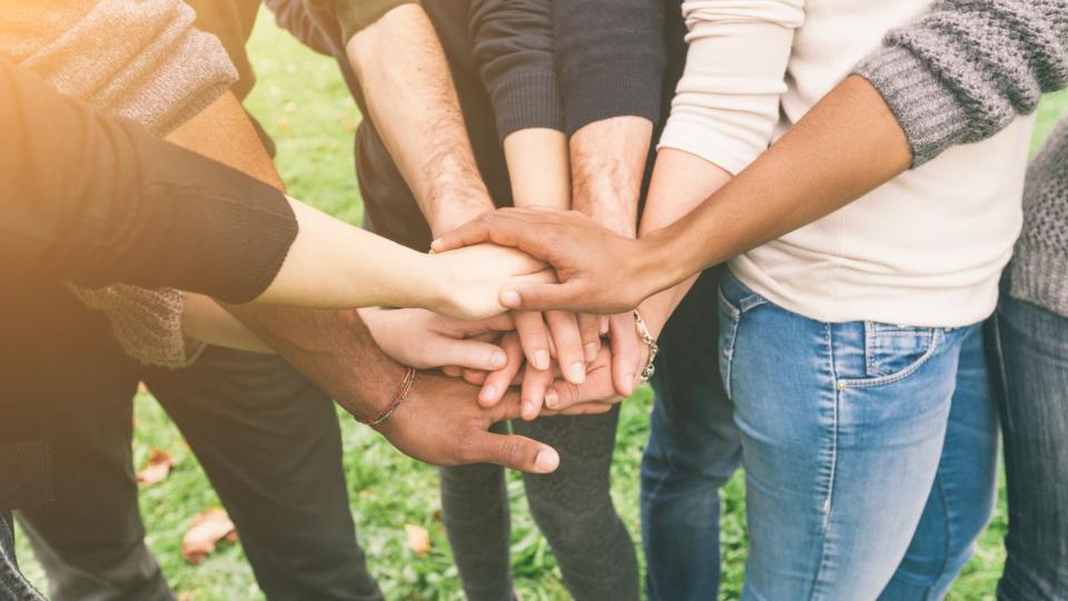 Abgebildet sind mehrere Personen unterschiedlicher Hautfarbe, die jeweils eine Hand in der Mitte des geformten Halbkreises übereinanderlegen. 