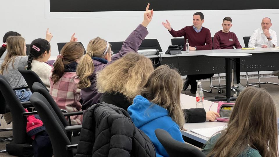 Abgebildet ist ein Seminarraum, in dem sich Schülerinnen und Schüler sowie Seminarleiter befinden. Einige Schülerinnen zeigen auf und ein Seminarleiter zeigt auf die Schülerin, welche die Antwort geben soll. 