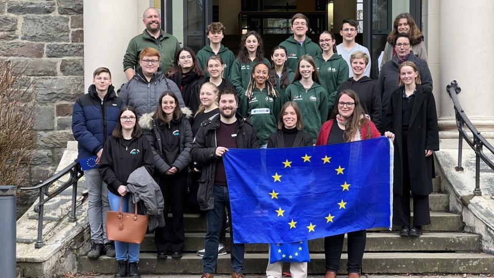 Abgebildet sind mehrere Personen, die gemeinsam auf einer Treppe für ein Bild posieren. Die Personen in der untersten Reihe halten eine Flagge der Europäischen Union. 