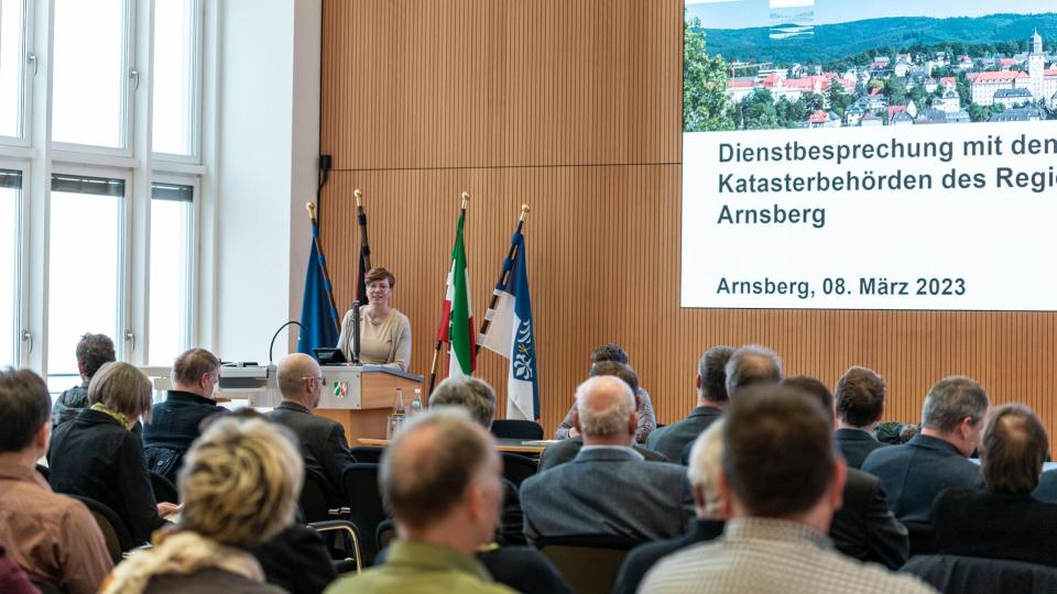 Abgebildet sind mehrere Personen, die sich in einem Sitzungssaal befinden. Eine Rednerin steht an einem Pult und andere Personen sitzen ihr gegenüber. 