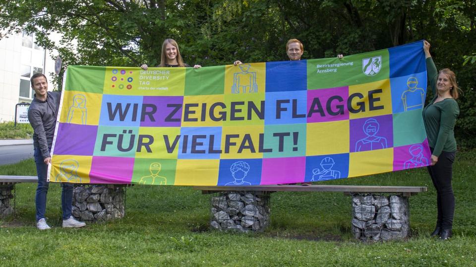 Vier Mitarbeiterinnen und Mitarbeiter der Bezirksregierung Arnsberg halten eine Flagge hoch. Die Flagge beinhaltet viele unterschiedliche Farben. Es steht geschrieben: "Wir zeigen Flagge für Vielfalt!".
