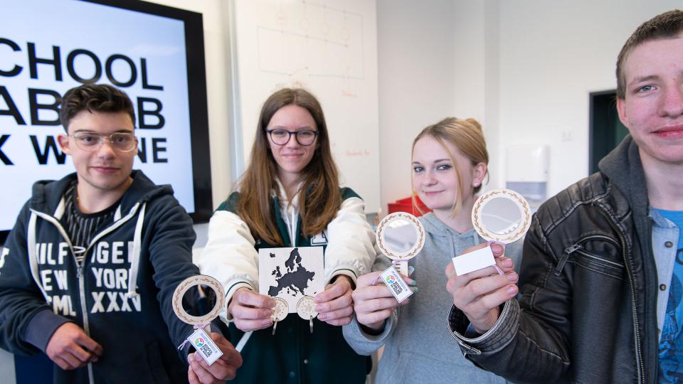 Abgebildet sind vier junge Erwachsene, die Schlüsselanhänger mit dem Logo von Patent im Handwerk, Reisespiegel mit einem Kamm als Griff und ein Schlüsselbrett mit einer applizierten Europa-Silhouette in den Händen halten.