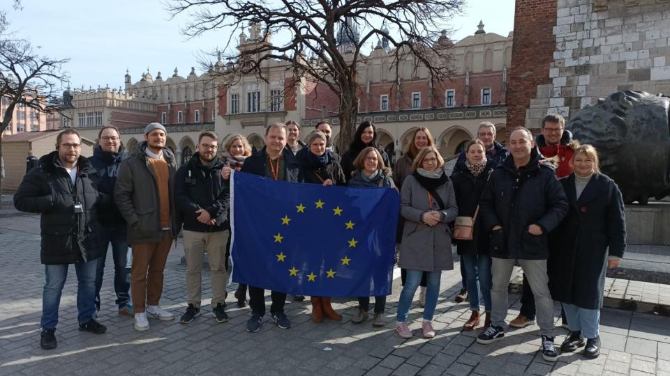 Abgebildet sind 26 Lehrkräfte der beruflichen Bildung, die vor einem imposanten Gebäude stehen und eine Flagge der EU hochhalten. 