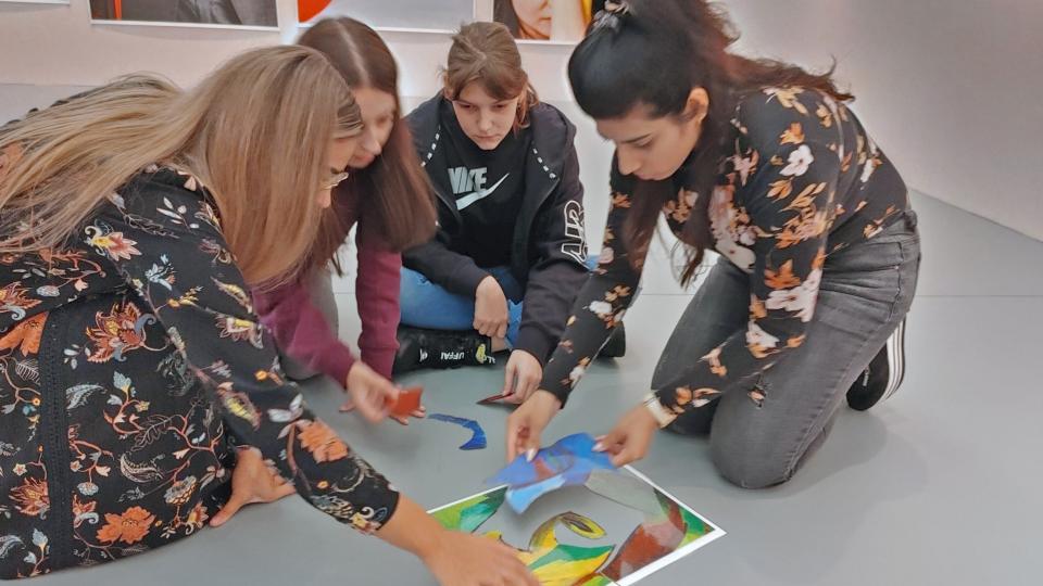Vier Studentinnen knien auf dem Boden in einem Museumssaal. Sie legen farbige Bildelemente zu einer Collage zusammen. An den Wänden im Hintergrund Ausschnitte von modernen Kunstwerken.