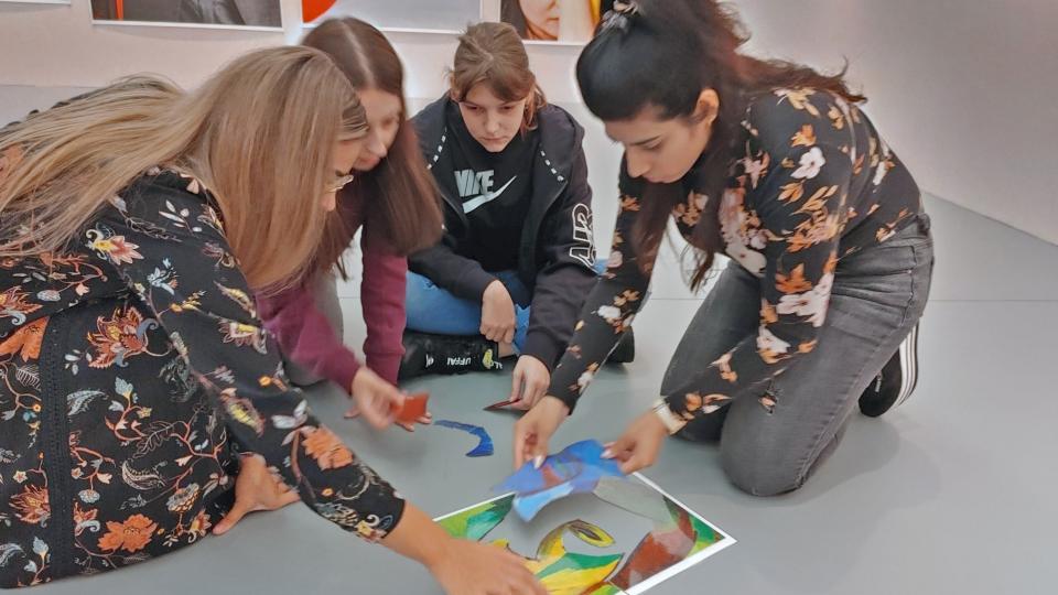 Vier Studentinnen knien auf dem Boden in einem Museumssaal. Sie legen farbige Bildelemente zu einer Collage zusammen. An den Wänden im Hintergrund Ausschnitte von modernen Kunstwerken. 