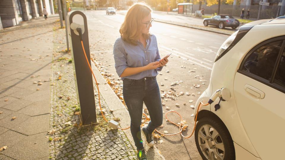 Junge Frau steht neben dem Elektroauto und schaut auf ihr Smartphone. Das Mietauto wird an der Ladestation für Elektrofahrzeuge aufgeladen.