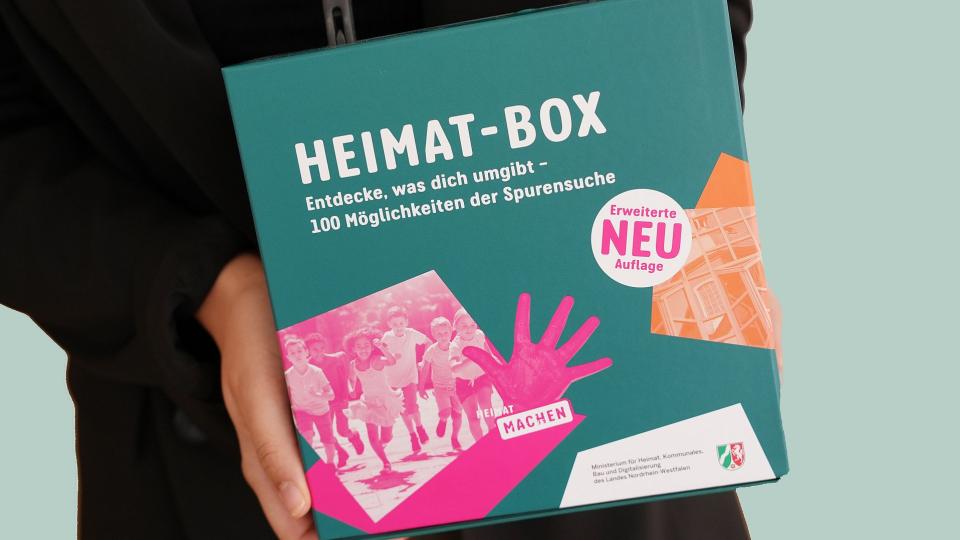 Abgebildet ist eine Person, die eine Box mit der Aufschrift "Heimat-Box" in den Händen hält.