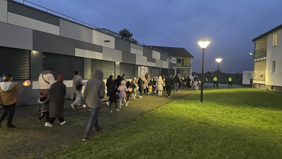 Mehrere Personen, die Kinder mit Laternen gehen an einem Gebäude vorbei. Zwei Straßenlampen leuchten.