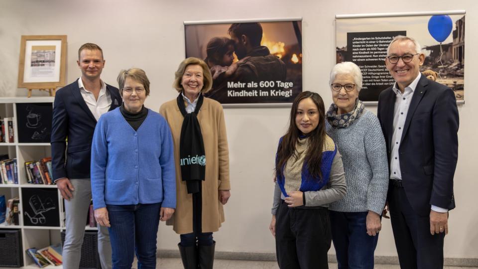 Gruppenbild von sechs Personen. Im Hintergrund hängt ein Poster mit der Aufschrift "Mehr als 600 Tage Kindheit im Krieg".