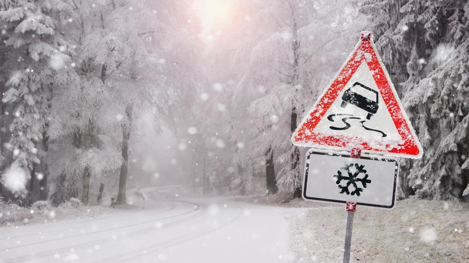 Eine Straße durch einen Wald. Es schneit stark. Im Vordergrund steht ein Warnschild, welches auf gefährliche Streckenführung hinweist.