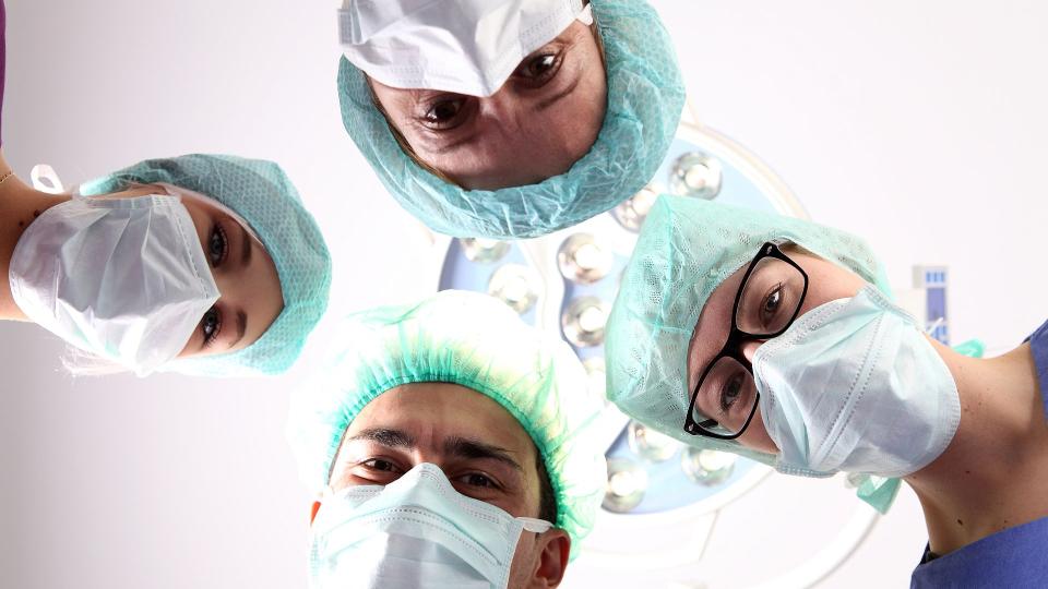 Abgebildet sind vier Anästhesietechnische Assistenzen beziehungsweise Operationstechnische Assistenzen, die in einem Operationssaal auf die zu behandelnde Person blicken.