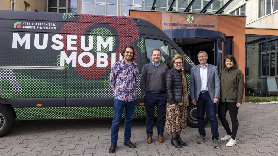 Fünf Personen stehen vor einem Transporter mit der Beschriftung Museum Mobil vor dem Haupteingang der Bezirksregierung.