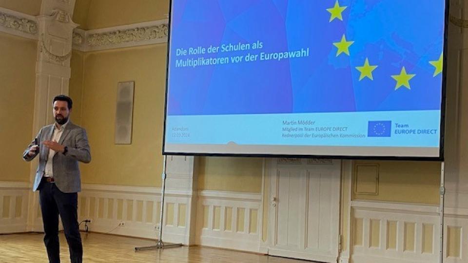 Abgebildet ist eine Person, die sich auf einer Bühne in einer Aula befindet und eine Rede zu dem Thema "Die Rolle der Schulen als Multiplikatoren vor der Europawahl" hält.