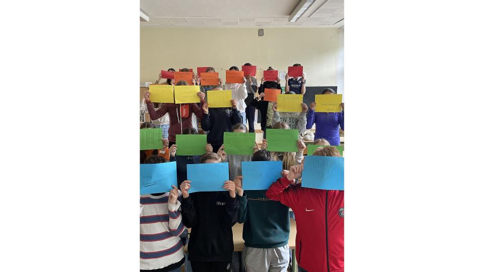 Abgebildet sind mehrere Kinder, die beschriftete bunte Blätter hochhalten.