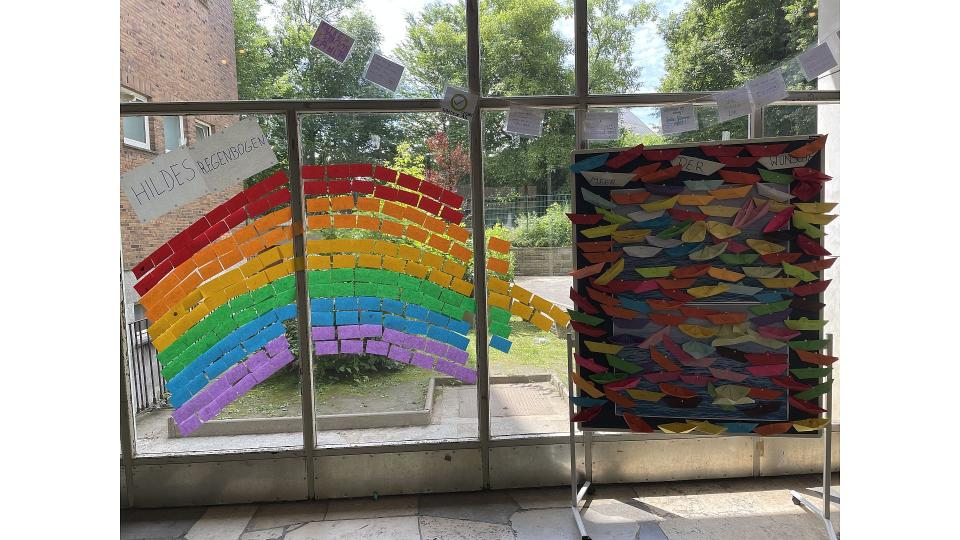Abgebildet sind bunte Zettel, die an eine Scheibe geklebt wurden. Die Zettel sollen in Farbe und Form einen Regenbogen darstellen. 