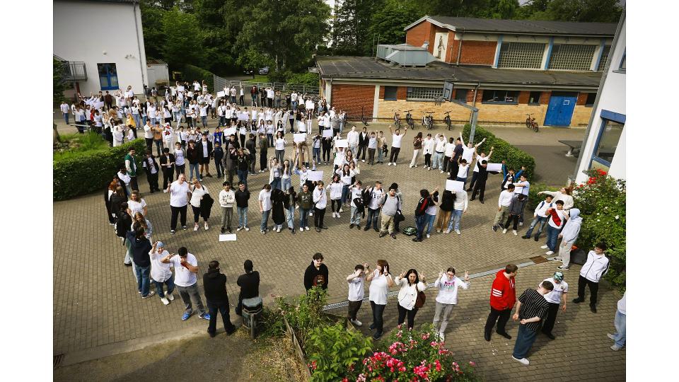 Abgebildet sind sehr viele Personen, die aufgereiht auf einem Schulhof stehen. 