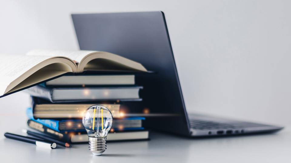 Abgebildet sind mehrere übereinander gestapelte Bücher, ein Laptop und eine angeschaltete Glühbirne, die im Fokus steht. 