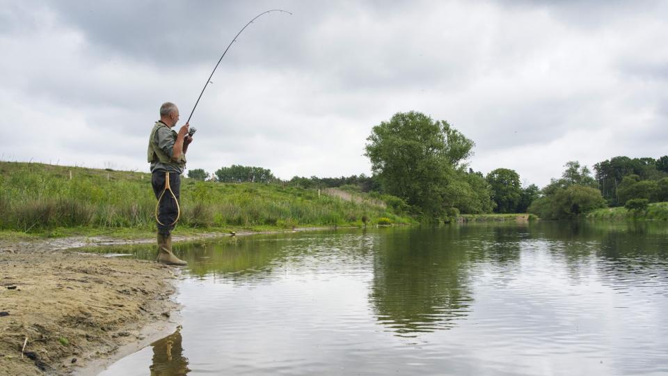 Abgebildet ist ein Angler, der an einem Fluss angelt.
