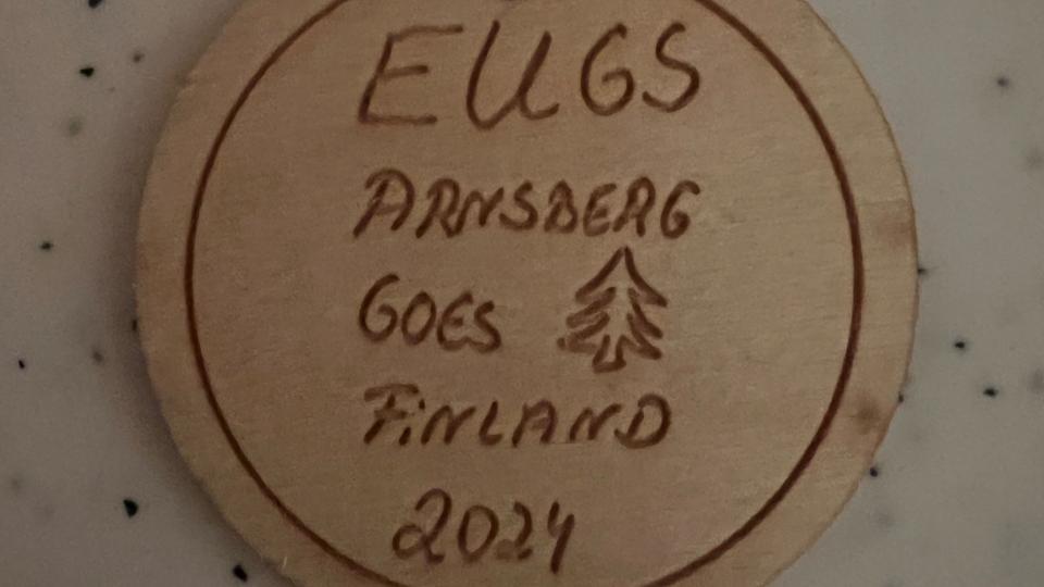 Abgebildet ist ein Holzuntersetzer mit der Aufschrift "EUGS Arnsberg goes Finland 2024".