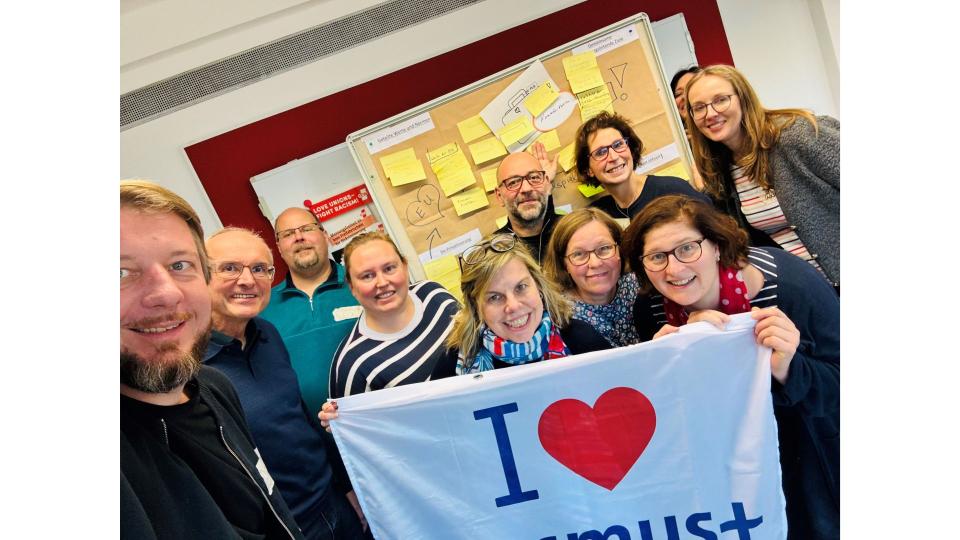 Abgebildet sind mehrere Personen, die für ein Bild posieren. Sie halten eine Flagge mit der Aufschrift "I love Erasmus+" (zu Deutsch: "Ich liebe Erasmus+") hoch.