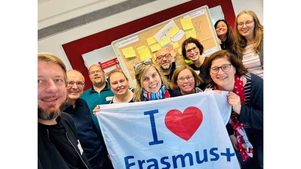 Abgebildet sind mehrere Personen, die für ein Bild posieren. Sie halten eine Flagge mit der Aufschrift "I love Erasmus+" (zu Deutsch: "Ich liebe Erasmus+") hoch.