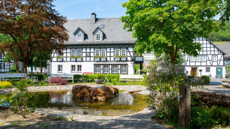 Abgebildet ist ein Fachwerkhaus in idyllischer Umgebung. Im Vorgarten des Hauses befindet sich ein Teich.