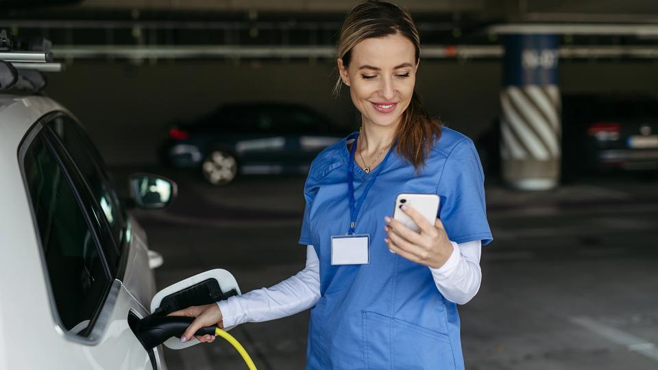 Abgebildet ist eine Pflegefachkraft, die ein Elektroauto in einer Tiefgarage lädt und gleichzeitig auf ihr Handy schaut.