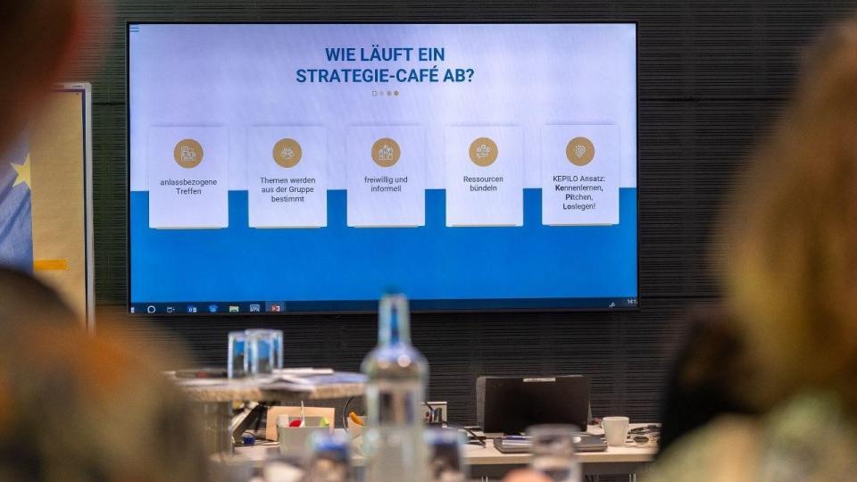 Abgebildet ist ein großer Fernseher in einem Schulungsraum, auf dem die Präsentation des Strategie-Cafés dargestellt wird.