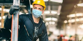 Abgebildet ist ein Mann in Arbeits- beziehungsweise Schutzkleidung, der sich in einer großen Fabrikhalle befindet. Der Mann trägt darüber hinaus eine Mund- und Nasenschutzmaske.