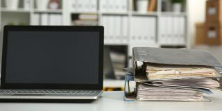 Ein aufgeklappter Laptop und zwei übereinandergestapelte Aktenordner auf einen Schreibtisch.