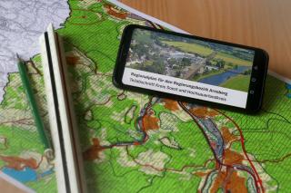 Abgebildet ist ein Handy, auf dessen Bildschirm "Änderungen des Regionalplanes Arnsberg – Teilabschnitt Kreis Soest und Hochsauerlandkreis" steht. Unter dem Handy befindet sich eine topographische Karte. 