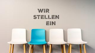 Vier Stühle, drei Weiße und einer in der Farbe Azur. Dieser ist beschriftet mit "Wir stellen ein"