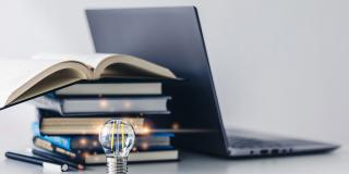 Abgebildet sind mehrere übereinander gestapelte Bücher, ein Laptop und eine angeschaltete Glühbirne, die im Fokus steht. 