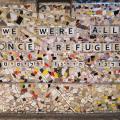 Ein Gemälde aus weißem und buntem Mosaikglas, auf dem "we were all once refugees" (zu Deutsch: Wir waren alle einmal Flüchtlinge) steht.