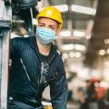 Abgebildet ist ein Mann in Arbeits- beziehungsweise Schutzkleidung, der sich in einer großen Fabrikhalle befindet. Der Mann trägt darüber hinaus eine Mund- und Nasenschutzmaske.