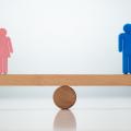 Auf der Balkenwippe aus Holz steht ganz links eine weibliche und ganz rechts eine männliche Figur. Die Wippe befindet sich im Gleichgewicht.