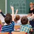 Mehrere Grundschulkinder befinden sich in einem Klassenraum und machen ein Handzeichen, welches andeutet, dass sie der an der Tafel stehenden Lehrerin etwas sagen wollen. 