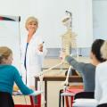Ein Gespräch zwischen einer medizinisch ausgebildeten Lehrerin und einem Schüler. In dem Klassenraum sind zwei weitere Schüler*innen sowie ein menschliches Skelett-Modell, das sich neben der Lehrkraft befindet, sichtbar.