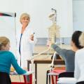 Ein Gespräch zwischen einer medizinisch ausgebildeten Lehrerin und einem Schüler. In dem Klassenraum sind zwei weitere Schüler*innen sowie ein menschliches Skelett-Modell, das sich neben der Lehrkraft befindet, sichtbar.