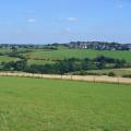 Panorama grüner Wiesen und einer eingezäunten landwirtschaftlich genutzten Fläche. Am Horizont befinden sich mehrere Häuser eines Dorfes.  