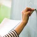 Eine Lehrerin steht vor einer Tafel. In der linken Hand hält sie ein Blatt Papier und mit der rechten Hand schreibt sie mit Kreide an die Tafel. 