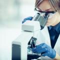 Eine Frau in einem weißen Kittel schaut durch eine Schutzbrille hindurch in eine großes Mikroskop