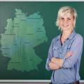 Eine Lehrerin steht vor einer grünen Tafel in einem Klassenraum auf der Tafel ist eine Karte von Deutschland eingeblendet, auf der alle Bundesländer zu sehen sind.
