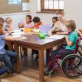 Ein Klassenraum mit mehreren Schülern an einem Tisch. Eine Lehrerein hilft einem Kind im Rollstuhl