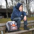 Ein Schüler sitzt alleine und nachdenklich auf einer Parkbank im Freien. Seinen Schulranzen hat er neben sich abgelegt. 